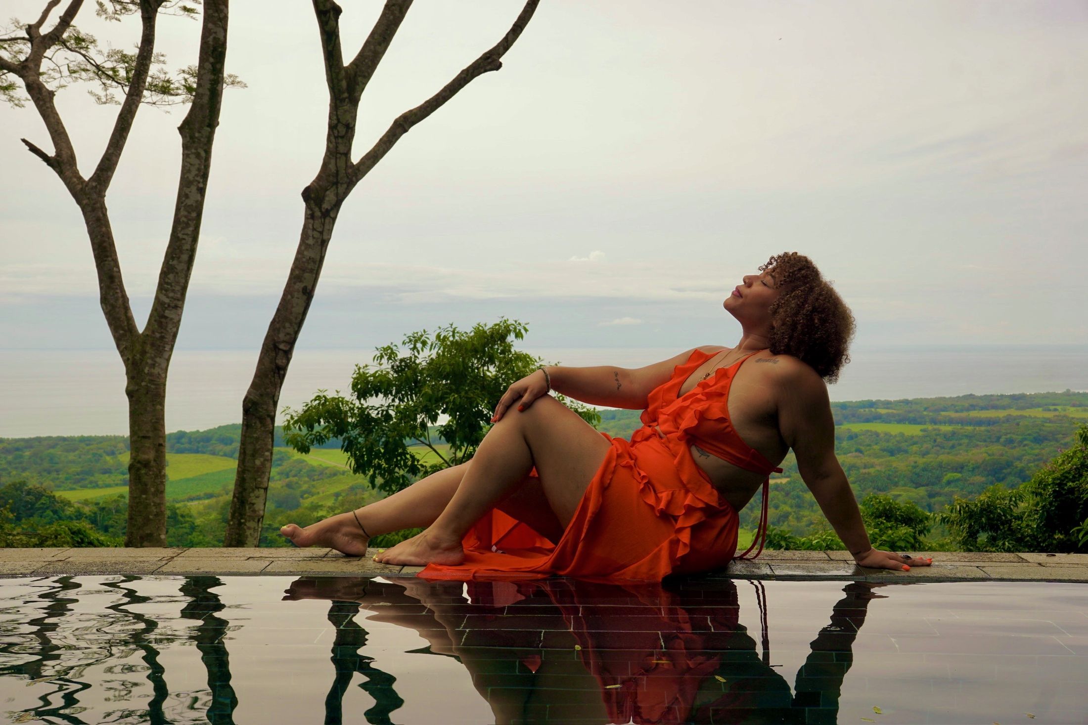 Danielle at the edge of an infinity pool in an orange dress, face tilted toward the sky, green hills and open water behind her.